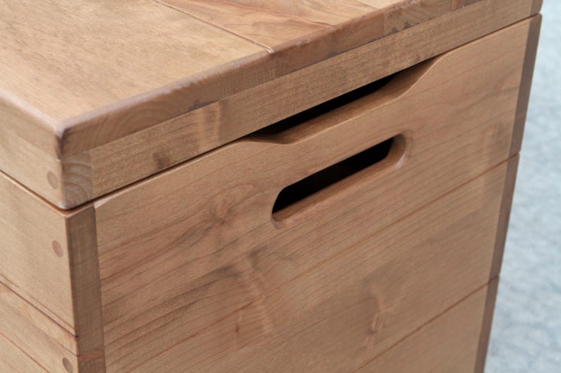 May include: Close-up of a wooden storage box with a natural finish. The box features a recessed handle for easy opening and closing. The wood grain is visible, and the box appears to be well-crafted.