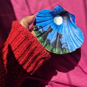 May include: A seashell painted with a blue night sky, a white moon, and black silhouettes of cats. The seashell is held in a hand wearing a red knitted sweater.