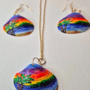 May include: A set of colorful shell jewelry featuring a sunset scene with palm trees painted on each piece. The set includes a necklace and a pair of earrings.
