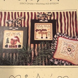 Könnte beinhalten: Ein weihnachtliches Kreuzstichmuster von Lizzie Kate. Das Bild zeigt gerahmte Stickmuster mit festlichen Designs, darunter ein Bär, ein Elch und die Worte "Peace, Joy, Love".