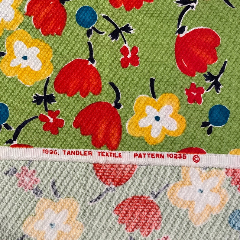 May include: A green fabric with a repeating pattern of red, yellow, and white flowers. The fabric has a woven texture and the text "1996. TANDLER TEXTILE PATTERN 10235 &copy;" is printed on the bottom edge.