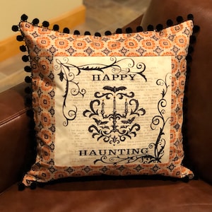 May include: A decorative cushion with a black and white print of a spooky mansion with bats, candles, and the text "HAPPY HAUNTING". The cushion has a black pom-pom trim and a patterned orange and black fabric backing.