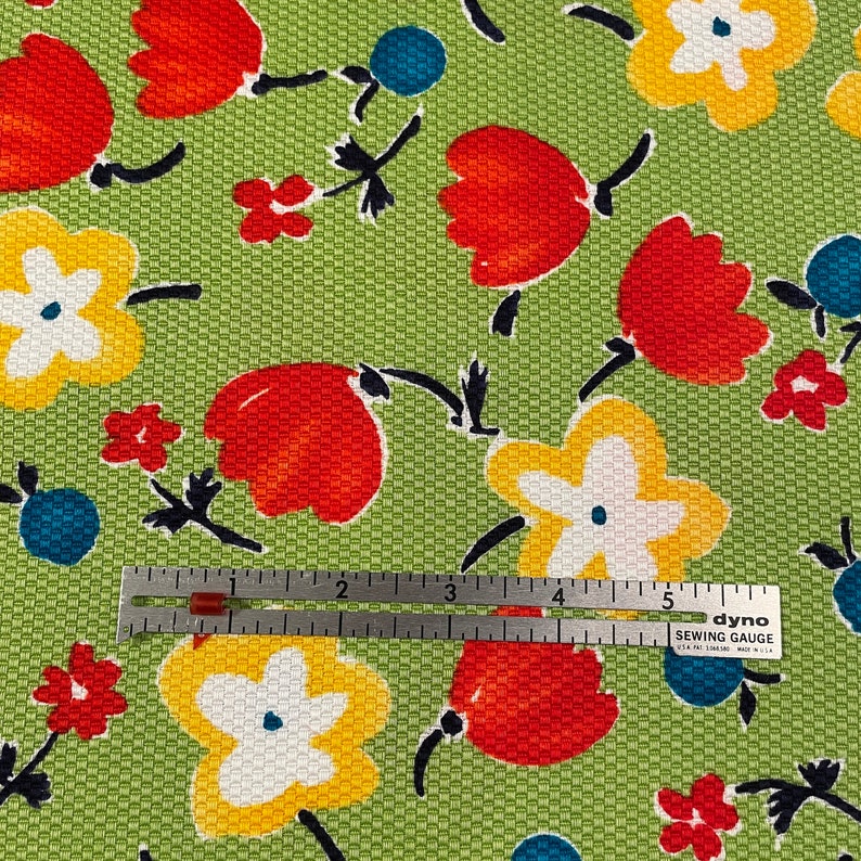May include: Green fabric with a repeating pattern of red, yellow, and white flowers. A ruler is placed on top of the fabric, showing measurements in inches.