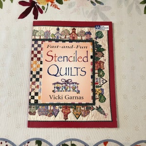 Puede incluir: Un libro titulado "Fast-and-Fun Stenciled Quilts" de Vicki Garnas. La portada presenta una ilustración colorida de una colcha con un patrón de casa y una cinta roja, blanca y azul.
