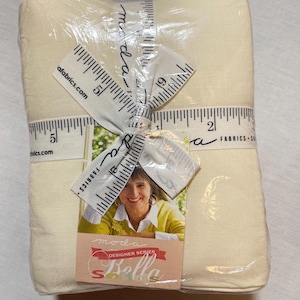Könnte beinhalten: Ein Bündel cremefarbener Stoffquadrate, verpackt in durchsichtigem Kunststoff. Ein weißes Band mit einem Lineal-Design und dem Wort "moda" umschließt den Stoff. Ein Etikett mit den Worten "moda DESIGNER SERIES Bella" und einem Foto einer Frau ist angebracht.