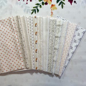May include: A collection of fabric bundles with various patterns, including floral, leaf, and text designs. The color palette is primarily white, cream, and orange, with accents of brown and gray. The fabrics are arranged on a white floral patterned surface.