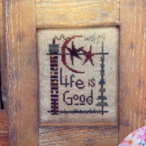 May include: A framed cross-stitch with the words "Life is Good" in black thread. The design includes a crescent moon, stars, and other stitched elements in red, green, and black. The frame is made of light brown wood.