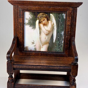 May include: A miniature wooden bench with a framed picture of a cherub. The bench is made of dark stained wood with a detailed design. The picture features a cherub with white wings and a soft, natural color palette.