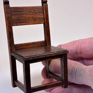 Può includere: Una sedia in miniatura di legno con finitura marrone scuro. La sedia ha un sedile rettangolare, uno schienale a doghe e quattro gambe quadrate. La sedia è tenuta da una mano, mostrando le sue piccole dimensioni e l'artigianato. Misure in centimetri.