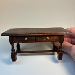 Op de afbeelding: Een miniatuur houten bureau met twee lades en messing knoppen. Het bureau heeft een donkerbruine afwerking en een rechthoekig blad. De poten zijn gedraaid en verbonden door een ligger. Het bureau is ongeveer 15 cm lang.