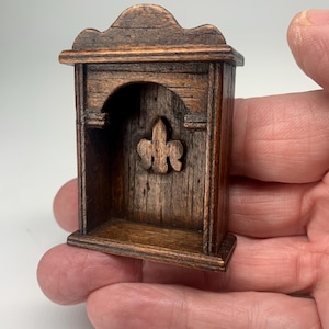Puede incluir: Un pequeño estante de madera marrón oscuro con un diseño de hoja tallada. El estante tiene una parte superior redondeada y una pequeña repisa. Es del tamaño de la mano de una persona.