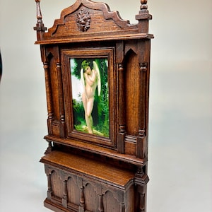 Puede incluir: Un mueble de madera en miniatura con una pintura enmarcada de un ángel desnudo en un bosque. El mueble, de color marrón oscuro, presenta tallas ornamentadas, incluida una cabeza de león. Tiene una repisa y columnas decorativas.