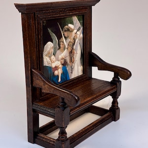 Può includere: Piccola sedia in legno marrone scuro con un'immagine religiosa incorniciata. La sedia ha braccioli e schienale a doghe. L'immagine raffigura angeli e figure in stile classico, con colori tenui e pennellate dettagliate.