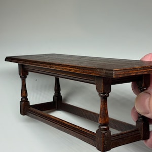 May include: A miniature wooden table with a dark brown finish. The table has a rectangular top and four turned legs. The table is approximately 15 cm long, 7.6 cm wide, and 7.6 cm tall.
