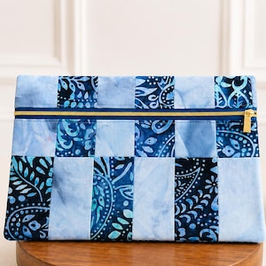 May include: A blue patchwork clutch with a gold zipper. The clutch features a variety of blue fabrics with floral and paisley patterns. The bag is rectangular and sits on a wooden stand.