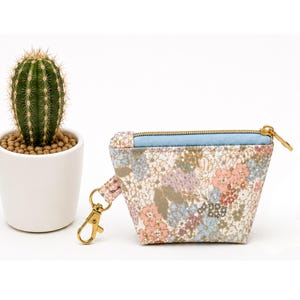 May include: A small fabric pouch featuring a multi-colored floral pattern in shades of pink, blue, and green. The pouch has a light blue top panel, a gold-toned metal zipper, and a gold-toned lobster clasp attached to the side with a fabric loop.