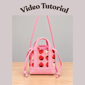 May include: A pink drawstring bag featuring a repeating strawberry pattern with red berries and green leaves. The bag has two short top handles and a long shoulder strap. The text 'Video Tutorial' is printed in dark brown above the bag.