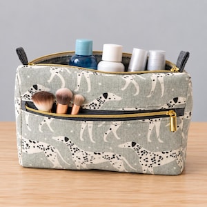 May include: A sage green rectangular toiletry bag featuring a repeating pattern of white and black spotted dogs. The bag has a gold-toned zipper closure, a front zippered pocket containing three makeup brushes, and four bottles stored inside.