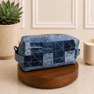 Puede incluir: Un neceser de patchwork de mezclilla azul con cierre de cremallera dorado y diseño acolchado. La bolsa rectangular presenta varios parches de mezclilla. Tiene asas pequeñas y se exhibe sobre un soporte de madera. Mide aproximadamente 25 cm de largo.