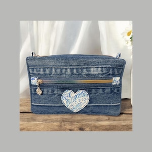 Pode incluir: Uma bolsa de ganga com fecho de correr dourado e um patch em forma de coração azul claro e branco. A bolsa tem um pequeno pendente floral. O fundo inclui uma superfície de madeira e tecido branco.