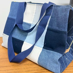 以下が含まれることがあります： パッチワークデニムのトートバッグ、青いナイロンストラップ付き。バッグは、明るいブルーと濃いブルー、青いストライプの白い生地など、さまざまなデニムの色合いが特徴です。バッグにはジッパー開閉部があります。