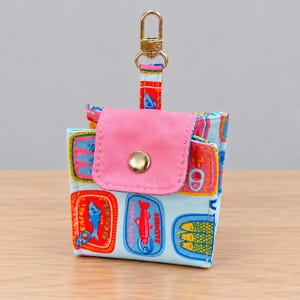May include: A small fabric pouch featuring a light blue background with a colorful sardine tin print. The pouch has a pink flap with a gold-tone snap closure and a gold-tone metal lobster clasp attached to a fabric loop at the top for hanging.