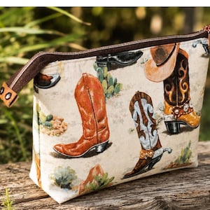Pode incluir: Uma bolsa com zíper feita de tecido cor de creme com uma estampa repetida de botas de cowboy marrons e brancas, cactos e paisagens desérticas. A bolsa possui um fecho de zíper marrom escuro com um pequeno puxador de metal.