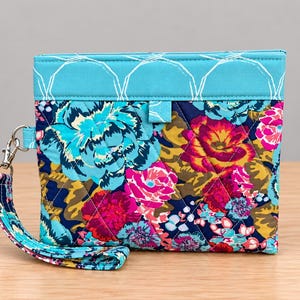 May include: A quilted wristlet pouch featuring a vibrant floral pattern with shades of turquoise, magenta, and navy blue. The top section is a solid turquoise fabric with white geometric stitching, and a matching wrist strap is attached with a metal clip.
