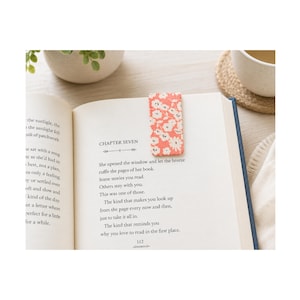 May include: A rectangular bookmark with a coral-colored background and a white floral pattern, placed inside an open book. The book displays printed text under the heading Chapter Seven, with visible page numbers and paragraph structures on the paper.