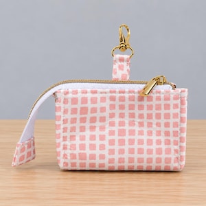 May include: A rectangular fabric pouch featuring a light pink grid pattern on a white background. The pouch includes a gold-toned metal zipper closure, a matching fabric pull tab, and a gold-toned swivel lobster clasp attached to a fabric loop.