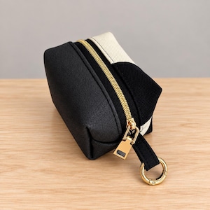 May include: A small rectangular pouch featuring a black textured exterior and a cream-colored canvas top panel. The pouch includes a gold-toned metal zipper closure with a matching gold ring pull attached to a black fabric tab on the side.