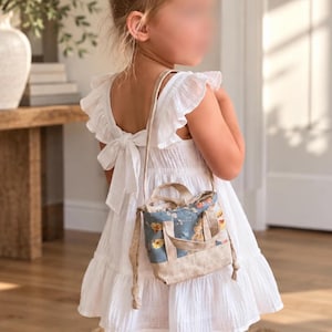 Könnte beinhalten: Ein weißes ärmelloses Kleid mit gerüschten Schultern und einer Schleife am Rücken, kombiniert mit einer kleinen Umhängetasche mit blauem Blumenmuster, gelben Blüten sowie beigefarbenen Stoffhenkeln und einem beigefarbenen Taschenboden.