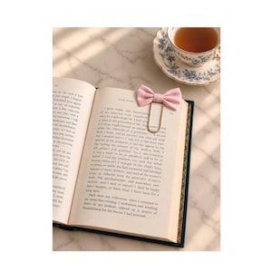 May include: A light pink fabric bow attached to a gold-toned metal paperclip bookmark, resting on the open pages of a book titled Jane Eyre, positioned next to a white and blue floral patterned teacup and saucer set on a marble surface.