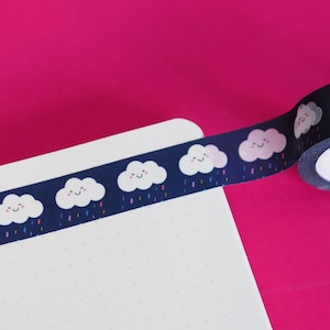 May include: A roll of washi tape with a repeating pattern of white clouds with smiling faces and colourful raindrops on a dark blue background. The tape is partially unrolled and is on a white surface with a dotted pattern.