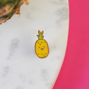 May include: A gold-coloured enamel pin shaped like a pineapple with a smiling face. The pineapple is yellow with green leaves and brown details.