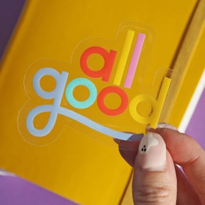 Könnte beinhalten: Ein transparenter Aufkleber mit den Worten "all good" in bunten Buchstaben. Die Buchstaben sind in einem Regenbogen-Farbschema gehalten, wobei das "a" rot, das "l" gelb, das "l" rosa, das "g" blau, das "o" türkis, das "o" rot und das "d" gelb ist.