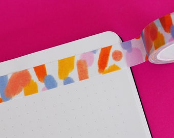 May include: A roll of decorative washi tape with a colorful abstract design of orange, yellow, blue, and pink shapes. The tape is applied to the edge of a white notebook with a dotted grid pattern, against a bright pink background.