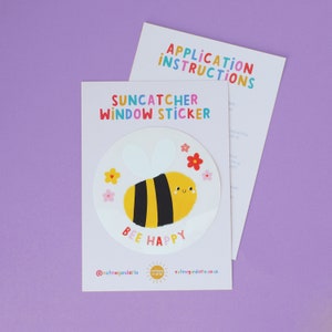 May include: A colourful window sticker with a cartoon bee design and the words "BEE HAPPY". The bee is yellow and black, surrounded by flowers. The packaging includes the text "Suncatcher Window Sticker" and "Application Instructions".