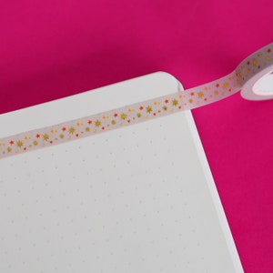Puede incluir: Un rollo de cinta adhesiva decorativa con estrellas doradas y corazones rojos se desenrolla sobre un cuaderno blanco punteado. La cinta es semitransparente y tiene un fondo rosa.