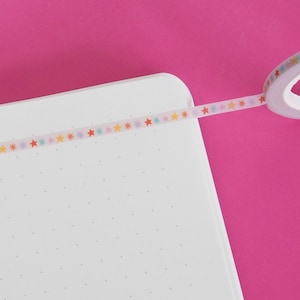 May include: A roll of decorative tape with colourful star patterns is partially unrolled over a white notebook. The tape features stars in various colours, including red, yellow, and blue. The background is a solid pink.