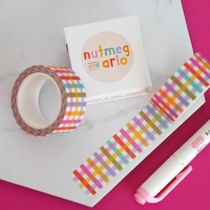 Könnte beinhalten: Eine Rolle buntes Gingham-Washi-Tape mit weißem Hintergrund. Das Band befindet sich in einer Schachtel mit dem Text "nutmeg and arlo" darauf.