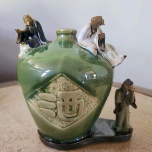 May include: A green ceramic vessel with a glossy finish, featuring three small sculpted figures attached to the exterior. A diamond-shaped panel on the front displays embossed Chinese characters. The vessel has a narrow neck and a dark base.