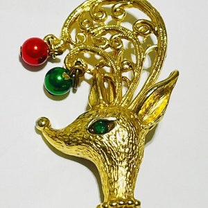 May include: Gold-toned reindeer brooch with a green and red beaded ornament hanging from its antlers. The reindeer has a green jewel for an eye.