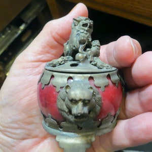 May include: A red and brass incense burner with a lion design. The burner has a round shape with a lid featuring two lion statues. The body of the burner has a lion's head on the front.