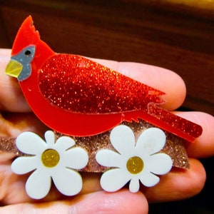 Peut inclure: Un cardinal rouge avec des accents de paillettes perché sur un lit de fleurs blanches avec des centres jaunes. Le cardinal est en plastique et est une pièce décorative.