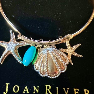 May include: A gold bangle bracelet with a seashell, two starfish, and a turquoise blue bead charm. The bracelet is on a black background with the text "JOAN RIVERS" visible at the bottom.