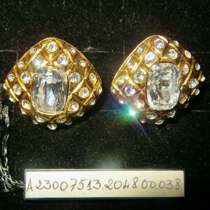 May include: A pair of gold-toned, diamond-shaped earrings with a large, clear, rectangular stone at the center. The earrings are encrusted with smaller, round, clear stones and set against a black background. A tag with the numbers A23007513.204800.038 is visible.