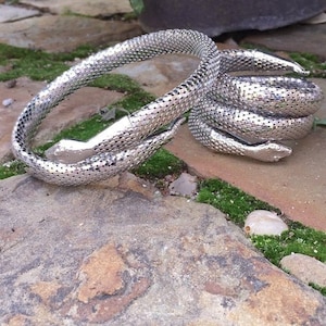 Peut inclure: Un bracelet en métal argenté en forme de serpent enroulé avec une surface texturée imitant des écailles. Ce modèle flexible présente une tête de serpent détaillée à une extrémité et une queue effilée à l'autre, formant une boucle circulaire.