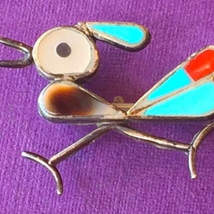 May include: A silver pin with a stylized bird design. The bird has a white shell body with black eyes, brown wings, and turquoise and red accents.