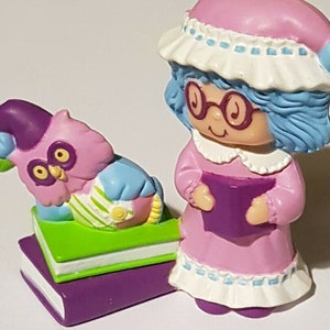 May include: A purple and blue owl figurine with a green and white book, sitting on a stack of purple books. A pink and blue figurine of a woman wearing a pink nightgown and glasses, holding a purple book.